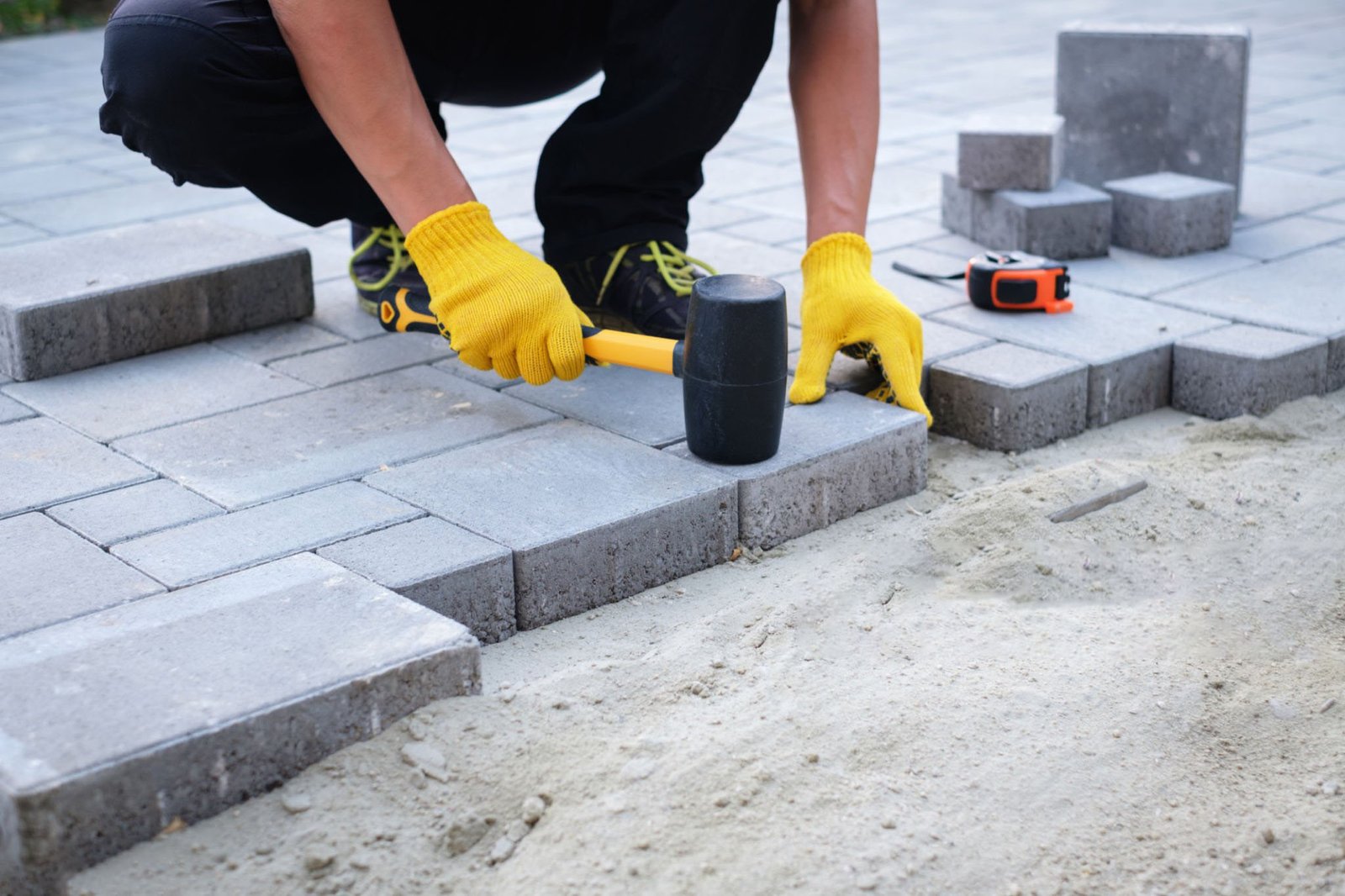 The master in yellow gloves lays paving stones in layers. Garden brick pathway paving by professional paver worker. Laying gray concrete paving slabs in house courtyard on sand foundation base.